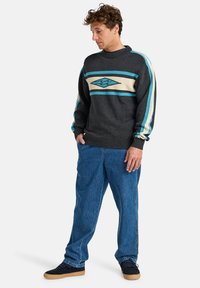 Gestrickter Pullover in Anthrazitgrau mit türkisfarbenen und cremefarbenen Streifenakzenten sowie einem Rautenmuster. Eine lockere Jeans in Blau und schwarze Sneakers.