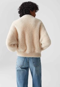 Person mit lockigem Haar, der eine cremefarbene, flauschige Jacke und eine blaue Jeans trägt, von hinten vor einem schlichten Hintergrund gezeigt.