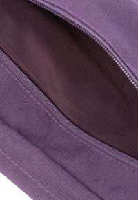 Lila Stofftasche mit Reißverschluss. Die Außenseite weist ein strukturiertes Muster auf, während das Innere glatt und dunkel lila ist.