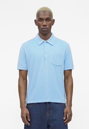 Homme aux cheveux courts décolorés porte un polo bleu clair à manches courtes avec poche avant et un jean bleu foncé sur fond blanc.