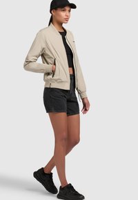 Helebeige bomber-jakk, millel on sile tekstuur, must tõmblukk ja soonilised varrukad, kombineeritud mustade teksashortsidega ja mustade tossudega.