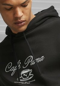 Black hoodie featuring white embroidered text "Café Puma" and a coffee cup graphic. Hood with drawstrings and soft fabric texture.