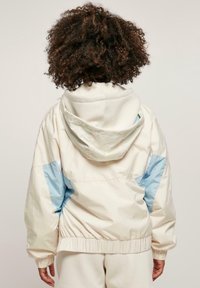 Chaqueta ligera con capucha en color crema y detalles en azul, con puños elásticos y dobladillo fruncido. Textura suave con un diseño casual.