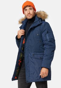 Parka en bleu marine isolée avec une capuche en fourrure, plusieurs poches et une doublure intérieure florale vibrante. Elle comporte une fermeture éclair.