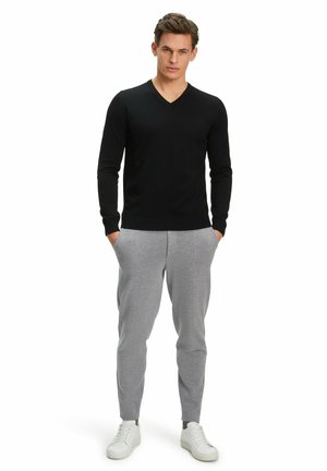 Zwarte V-hals trui van gebreid materiaal, gecombineerd met lichtgrijze tapered joggingbroek. Model draagt witte sneakers en heeft de handen in de zakken.