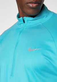 Ingrandimento di una persona che indossa una felpa sportiva azzurro chiaro con zip Nike, mostrando il petto e la parte bassa del viso.