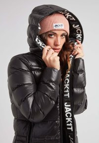 Mujer con una chaqueta acolchada negra con capucha y un gorro de punto rosa etiquetado "JACKIT", ajustando la capucha alrededor de su rostro.