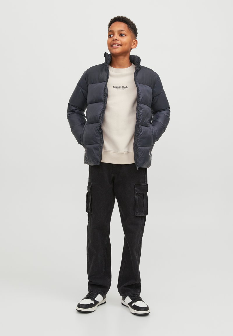 Črna puhovka z visokim ovratnikom, nosena čez svetlo majico, v kombinaciji z črnimi cargo hlačami. Obutev: bele in črne športne copate.