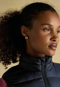 Melna pūkaina veste ar augstu apkakli un rāvējslēdzēju. Modelis ir ar cirtainiem matiem un valkā zelta krāsas kvadrātveida auskarus, fonā ir neitrāls attēls.