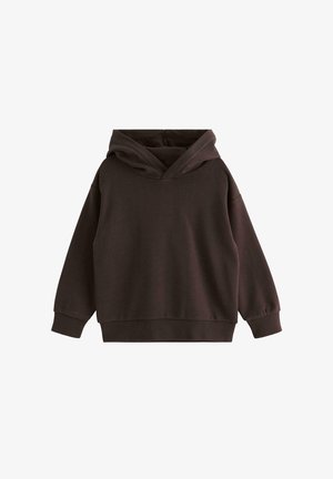 Smilende lille barn justerer mørkeblå strikhue med bjørnepatch, iført brun sweatshirt og olivengrønne bukser, foran ensfarvet baggrund.
