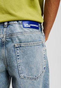 Jeans de mezclilla azul claro con una etiqueta de cuero azul en la cinturilla que dice "KARL LAGERFELD JEANS". Presenta bolsillos traseros clásicos y costuras.