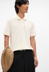 Μπεζ πόλο μπλουζάκι με κλασικό γιακά, κοντά μανίκια και χρυσό λογότυπο. Συνδυασμένο με μαύρο παντελόνι και καφέ πουλόβερ κρεμασμένο στον βραχίονα.