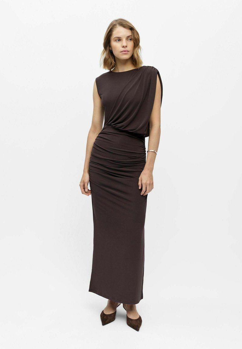 Femme portant une robe longue sans manches, marron foncé, froncée, avec un détail drapé asymétrique à l'épaule et des chaussures à bouts pointus assorties.