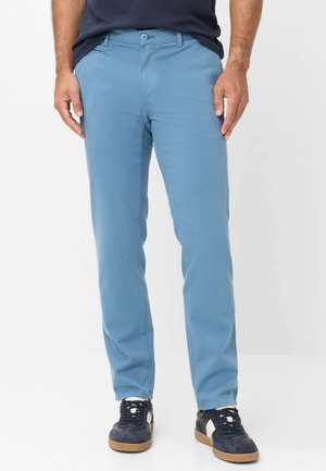 Mann trägt hellblaue gerade geschnittene Hose, navyblaues Hemd und navyblaue Sneakers mit weißen Akzenten vor einem schlichten weißen Hintergrund.