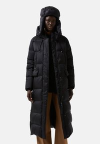 Manteau long noir en duvet avec capuche, doté d'une texture matelassée, d'une fermeture éclair à l'avant, de poches latérales et d'une silhouette à coupe large.