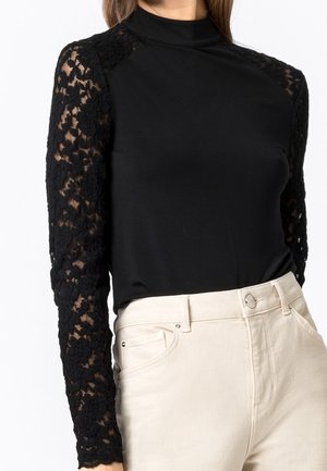 Femme portant un haut noir avec des manches longues en dentelle florale, associé à un pantalon beige clair taille haute, sur un fond uni.