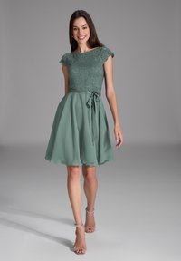 Grünes Kleid mit einem Spitze-Oberteil, kurzen Ärmeln und einer Schleife in der Taille. Fließender Rock mit weicher Textur und knielanger Saum.