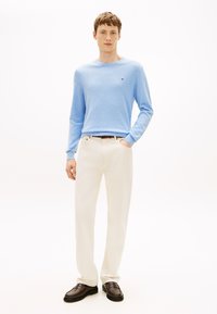 Světlo modra srajca z dolgimi rokavi v mehki teksturi, kombinirana s kremastimi hlačami in temno rjavimi loafer čevlji. Na srajci so minimalni logotipi.