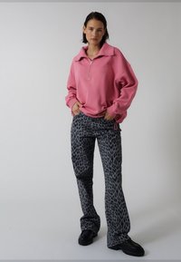 Różowa bluza zapinana na zamek z szerokim kołnierzem, zestawiona z szarymi spodniami w panterkę. Modelka ma na sobie czarne, masywne buty. Proste, neutralne tło.