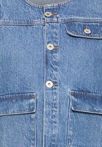 Nærbillede af blå denimjakke med fire metalknapper og tre frontlommer med klap, der er syet med orange syninger.