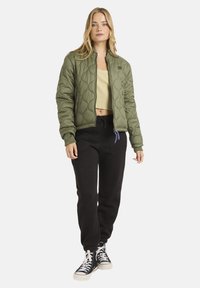 Ung kvinde iført en olivenfarvet quiltet jakke, beige cropped top, sorte jogger bukser og sorte høj-æble sneakers, stående mod en hvid baggrund.