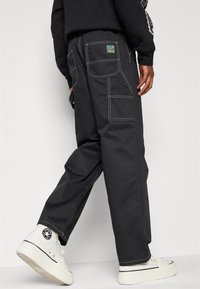 Czarne spodnie cargo wykonane z wytrzymałego materiału, z wieloma kieszeniami, kontrastującymi białymi szwami i luźnym krojem. W połączeniu z białymi sneakersami.