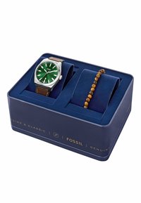 Reloj Fossil con esfera verde, caja plateada y correa de cuero marrón, en una caja azul marino. Incluye una pulsera con cuentas en tonos marrón y amarillo.