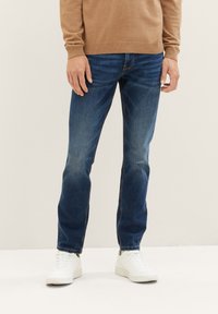 Hombre vestido con jeans azules, zapatillas blancas y un suéter marrón, de pie contra un fondo liso de color blanco roto.