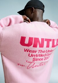 Oseba v roza puloverju z rdečim napisom "UNTITLED Wear The Unknown Untitled Goods Since 2001" in črnim klobukom, obrnjena stran od kamere.