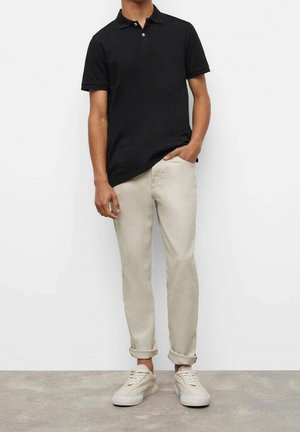 Mand iført en sort polo, lyse beige optrukne bukser og hvide sneakers, stående mod en ensfarvet hvid baggrund.