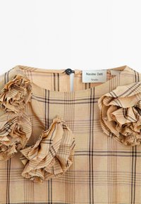 Beige geruite jurk met drie textuurstof ruches op elke schouder. Voorzien van een ronde halslijn en een knoopsluiting aan de achterkant.