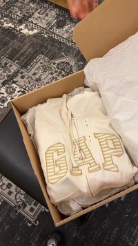 Hanorac cu fermoar GAP crem, pliat, într-o cutie de carton deschisă cu hârtie de ambalat albă, așezată pe o mochetă cu model. O mână ridică capacul cutiei.