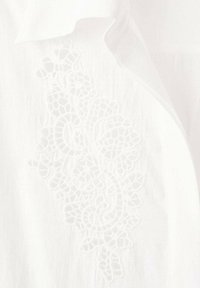 Blusa blanca con bordado blanco intrincado en un patrón floral en el lado izquierdo. Tela suave y ligera con una textura lisa.