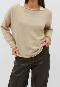 Beżowy sweter dzianinowy o luźnym kroju, szerokim dekolcie, ściągaczach i dolnym wykończeniu. W połączeniu z ciemnobrązowymi spodniami o gładkiej fakturze.