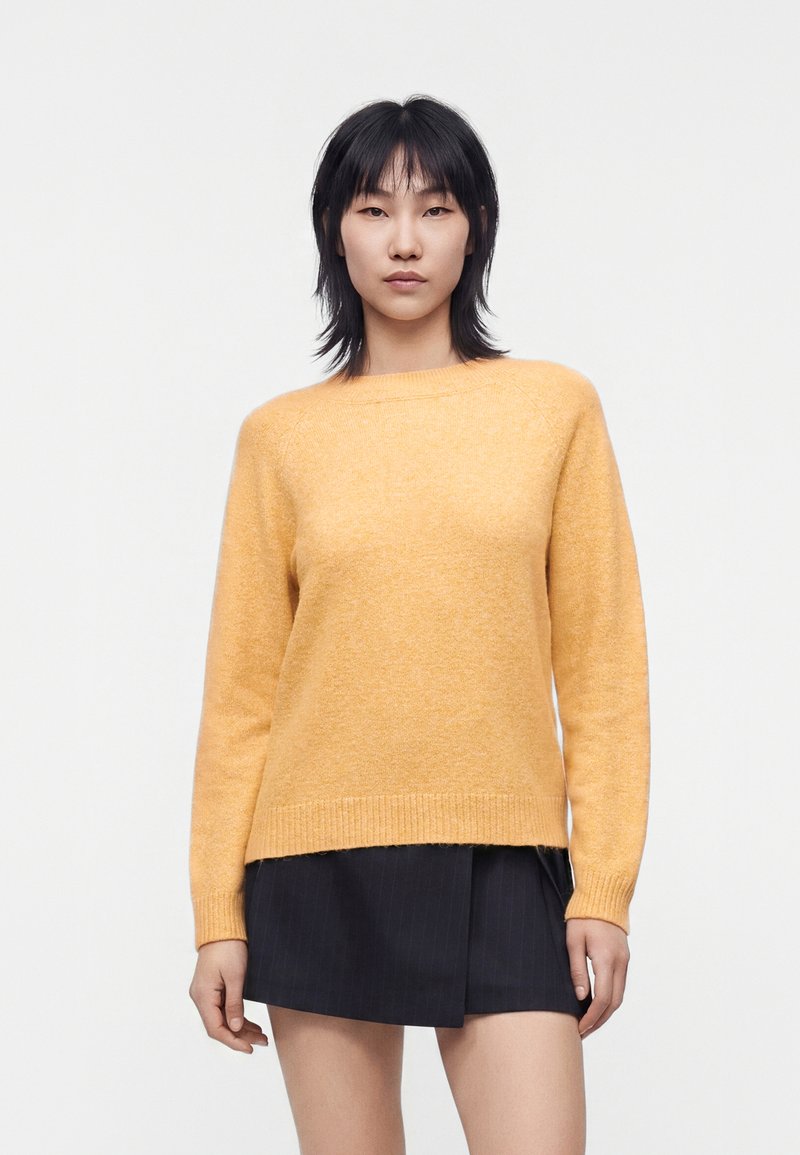 Kvinde med sort hår iført en sennepsgul sweater og en sort kort nederdel, stående mod en ensfarvet hvid baggrund.