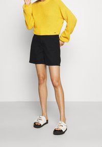 Suéter de punto amarillo, pantalones cortos negros y sandalias blancas con suelas negras. El suéter tiene un cuello redondo; los pantalones cortos son de longitud hasta la rodilla. Conjunto informal.