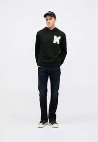 Felpa nera con cappuccio e un design grafico bianco sul petto, abbinata a jeans scuri e sneakers nere con suola bianca.