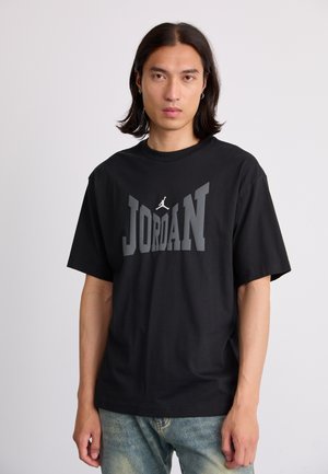 Black cotton t-shirt featuring bold "JORDAN" text in gray and white, crew neck, and short sleeves, paired with light denim jeans.