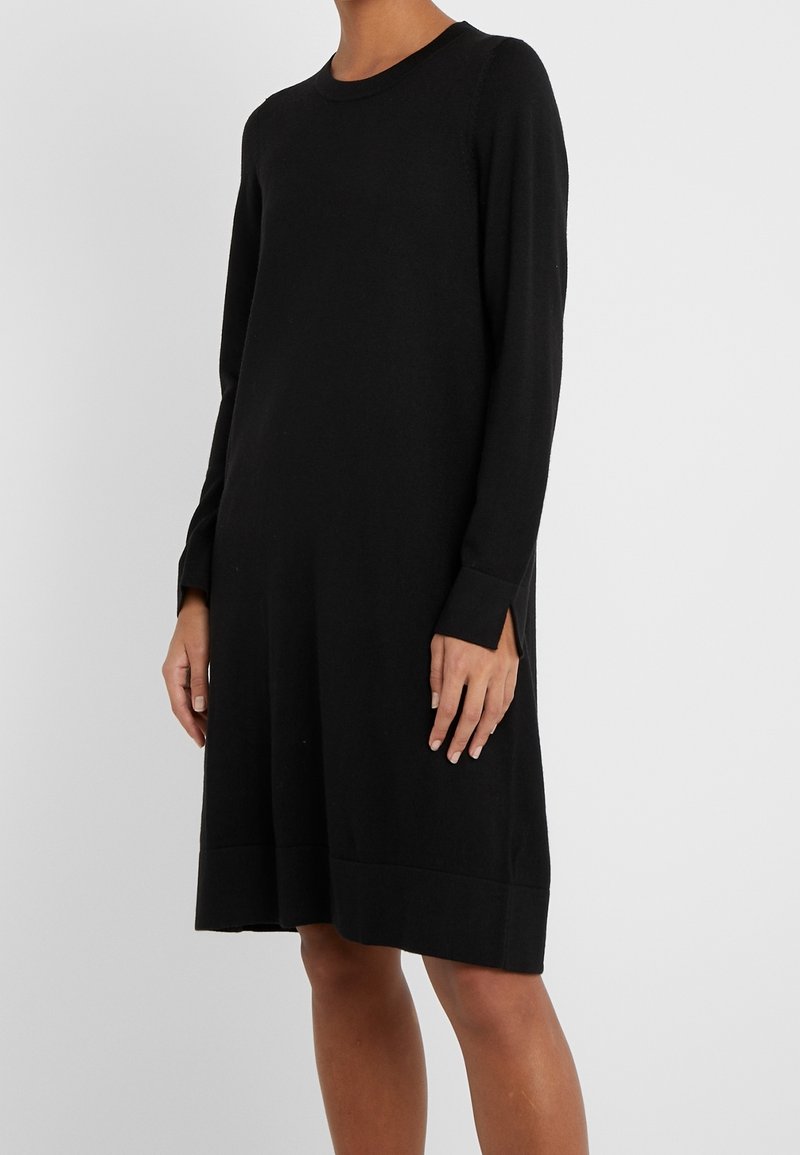 Personne portant une robe noire à manches longues, longueur genou, avec un col rond et de petites fentes au niveau des manches, debout devant un fond uni.