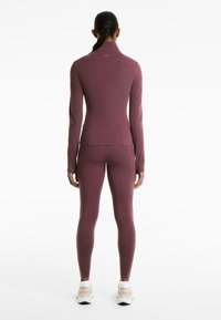 Femme se tenant de dos, portant un haut moulant mauve à manches longues et des leggings assortis, avec des chaussures de sport blanches, les cheveux en tresse.