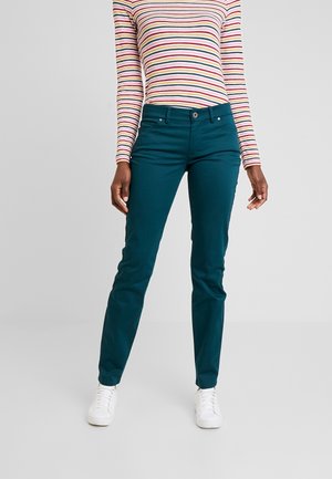 Kobieta ubrana w dopasowane spodnie w kolorze morskim, białe trampki i długi rękaw z wielokolorowymi paskami, stojąca na tle prostego jasnoszarego tła.