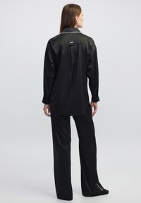 Camicia nera in satin con colletto, caratterizzata da una rifinitura decorativa lungo il retro, abbinata a pantaloni larghi coordinati.