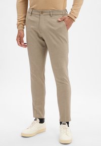 Pantalon beige sur mesure avec un devant plat, des poches latérales et une fermeture à un bouton, associé à des baskets blanches et des chaussettes noires.