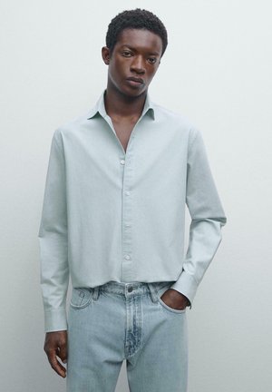 Young man with dark skin wearing a pale green long-sleeve button-up shirt and light blue denim jeans, one hand in pocket, neutral background.