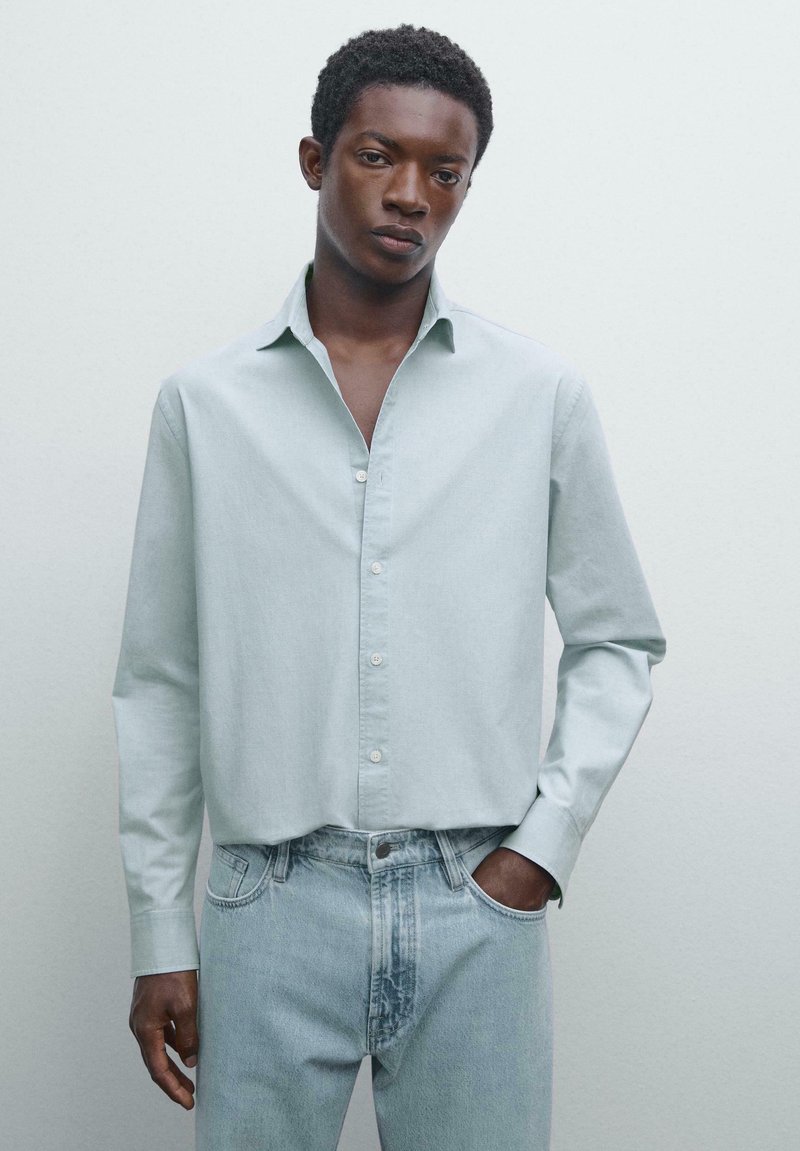 Giovane uomo con pelle scura che indossa una camicia a maniche lunghe verde chiaro con bottoni e jeans in denim blu chiaro, con una mano in tasca, sfondo neutro.