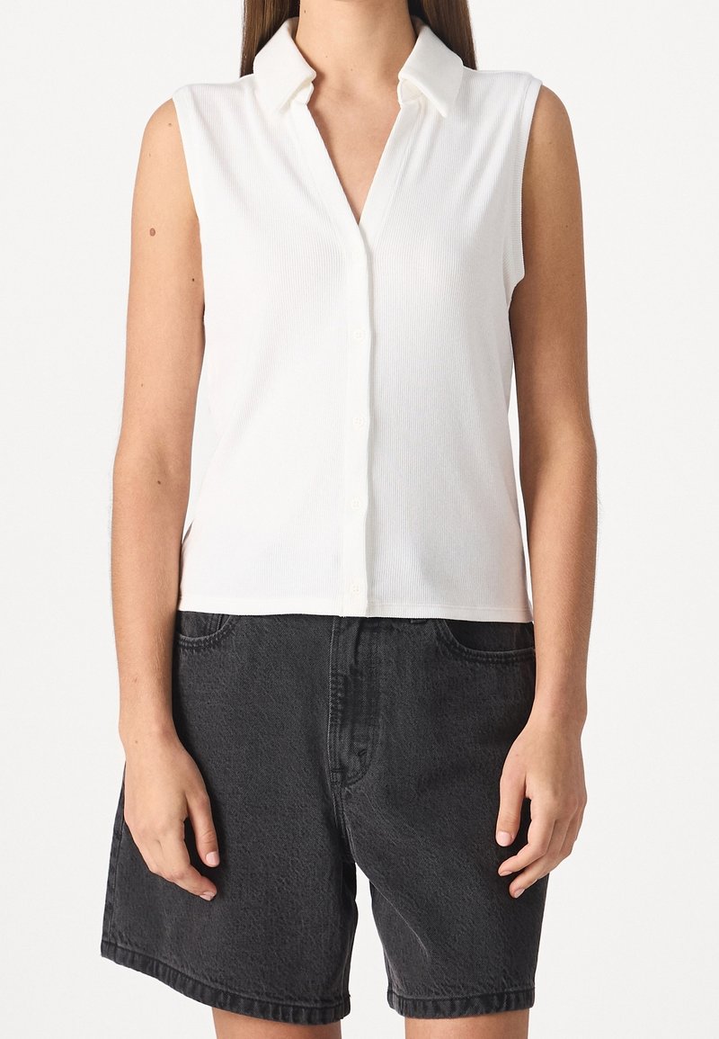 Femme portant une chemise sans manches blanche à boutons et un short en denim noir ample, se tenant devant un fond blanc uni.