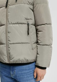 Leichtgraue Pufferjacke mit gesteppten Bereichen, Reißverschlusstaschen und einem schwarzen Rippbündchen. Über einer Jeanshose getragen.