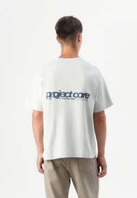 Una persona se encuentra de espaldas al espectador, vistiendo una camiseta gris claro con el texto "project core" en azul oscuro, combinada con pantalones beige.