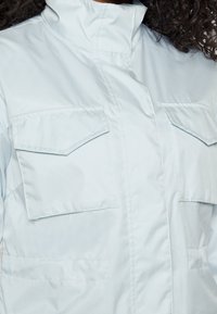 Ljusblå jacka med hög krage, två bröstfickor och en slät, glänsande yta med minimalistiska designelement.