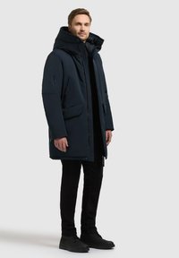 Parka imbottita blu navy con collo alto, tasche con zip e chiusure a scatto. Presenta un design elegante e moderno ed è realizzata in materiale resistente alle intemperie.